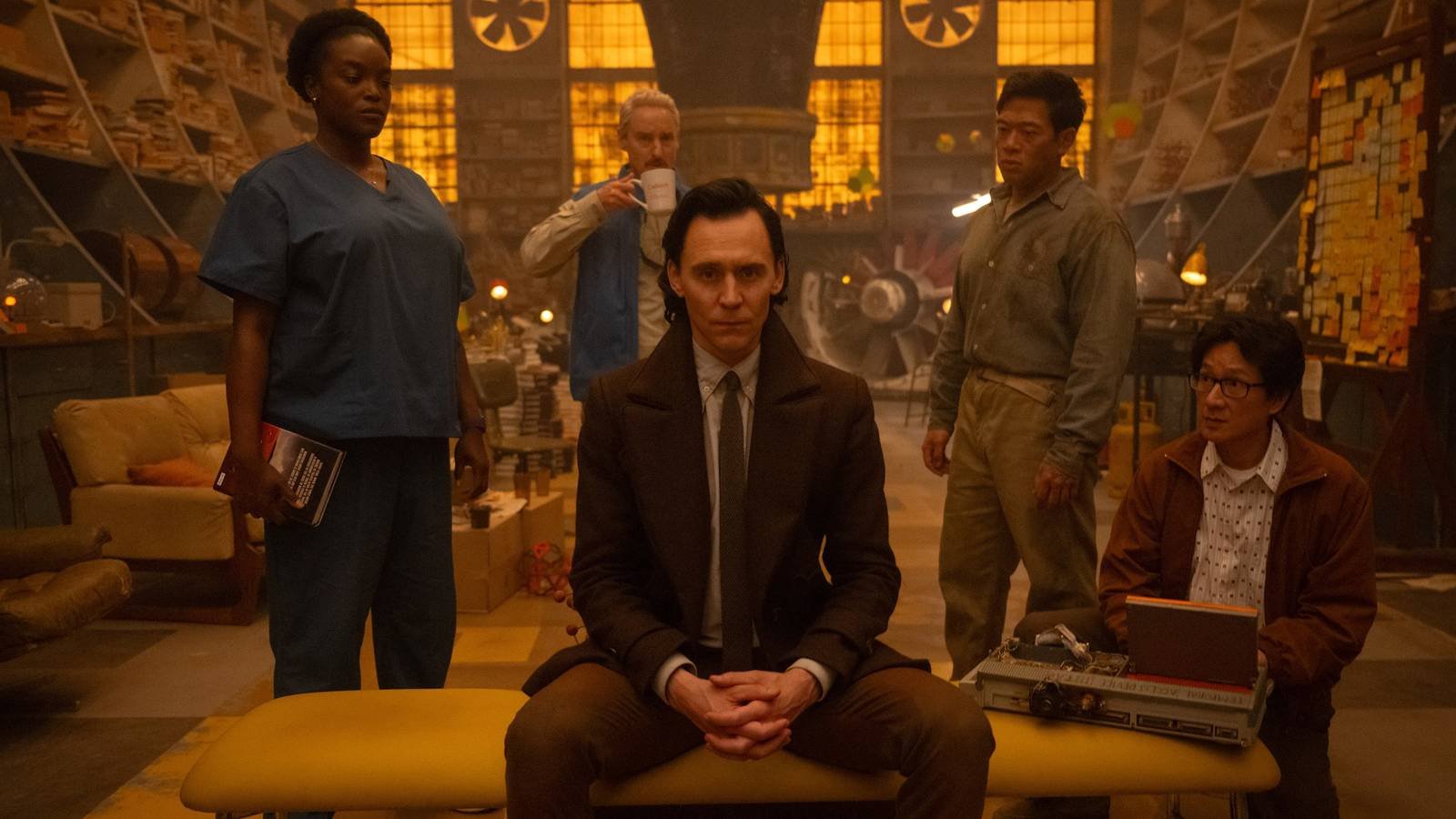 Loki (Tom Hiddleston) sitting facing the camera with supporting characters around him in Loki sea...